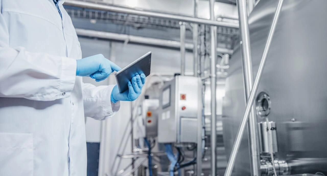 A worker in a lab coat and gloves is studying something on a tablet.