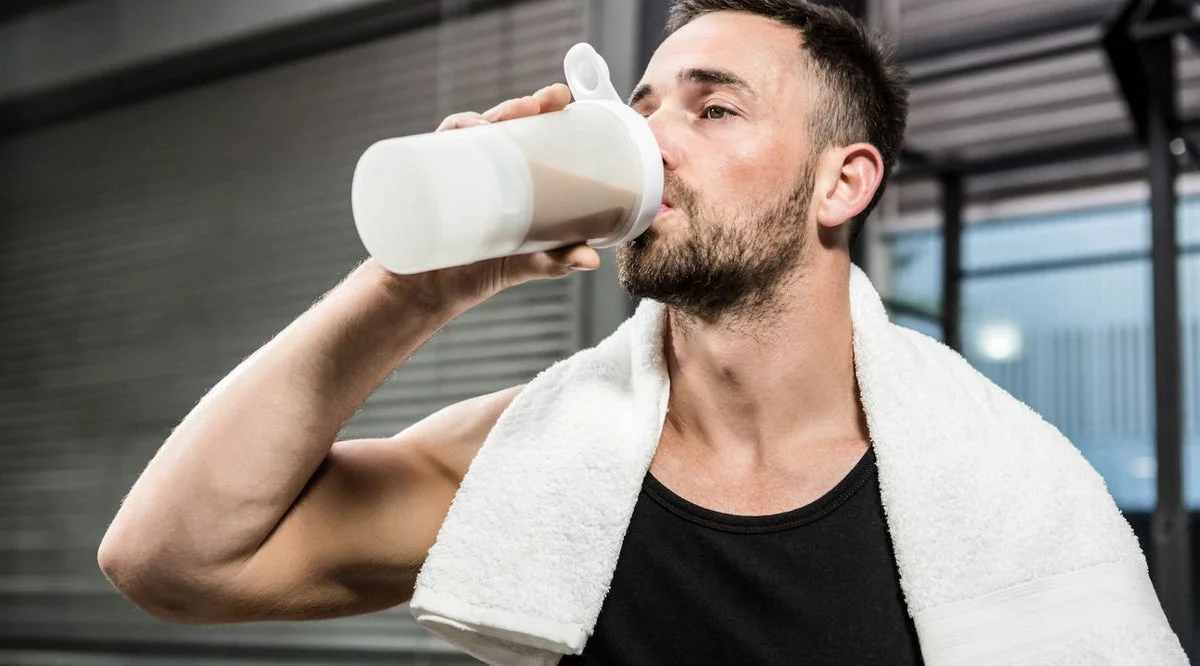 A man drinking a protein shake in a gym with a towel around his neck